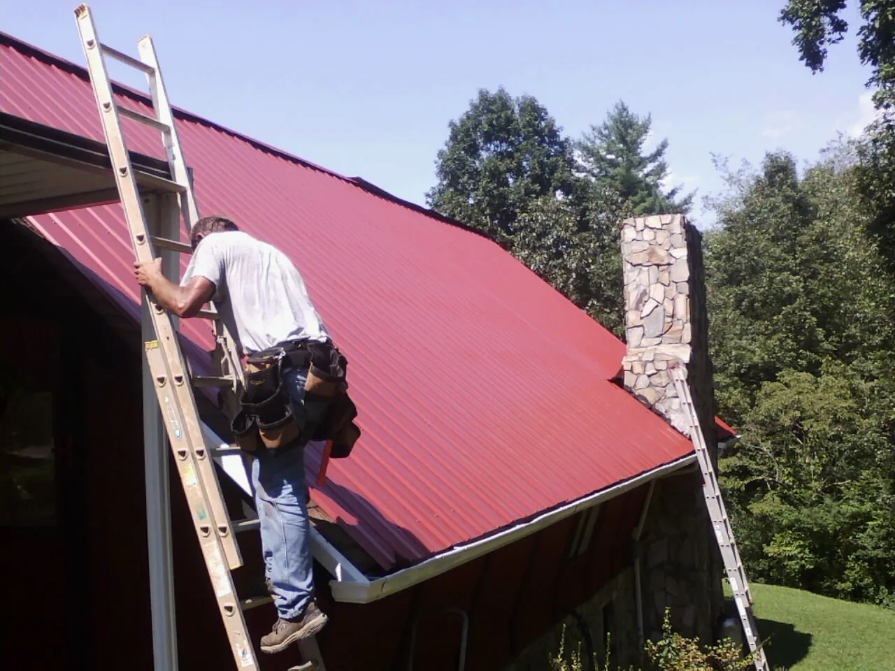 Roofer on a ladder beside a red metal roof and stone chimney — Soffit & Fascia Repair by Roofing Brothers in Byrdstown